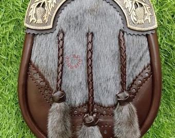 May include: A brown leather sporran with a gray fur front and decorative bronze thistle accents. The sporran features three braided leather tassels with gray fur ends, and is set against a green background.