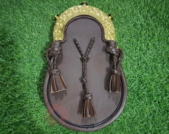 May include: A brown leather sporran featuring a polished gold-tone metal cantle with an embossed circular pattern. The front is decorated with three braided leather cords, each ending in a matching brown leather tassel hanging from the top.