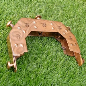 Puede incluir: Un objeto de metal de color bronce con un diseño curvo en forma de U. Presenta remaches decorativos y patrones circulares. El objeto tiene una estética vintage y está colocado sobre un fondo de hierba verde.