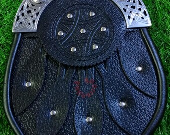Könnte beinhalten: Ein schwarzer Leder-Sporran mit silbernem keltischen Design auf der Klappe und Oberseite. Der Sporran hat ein kreisförmiges Design in der Mitte und dekorative silberne Nieten. Ein traditionelles schottisches Accessoire.