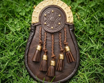 May include: A dark brown leather sporran featuring a polished brass cantle with decorative circular engravings and studs. The front flap displays a central circular leather emblem with brass accents and five braided leather tassels with brass caps.