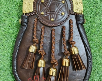 May include: A brown leather sporran with a decorative brass top and five tassels. The sporran features a circular design with a star pattern and brass studs. The tassels have brass caps and are attached to braided leather cords.