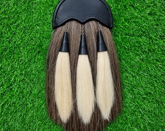 May include: A sporran featuring a black leather top flap with a curved edge. The body is made of long, brown fur, accented by three hanging white fur tassels, each attached to the top flap by a small metal chain and a black conical cap.
