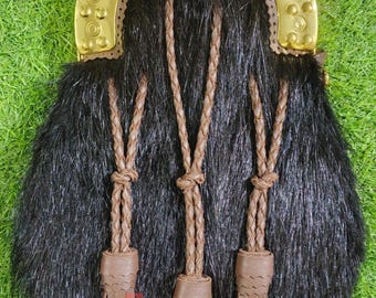 May include: A black fur sporran with a decorative brass top and brown leather accents. The sporran features three braided leather tassels with leather caps and fringe. The brass top has an ornate design.