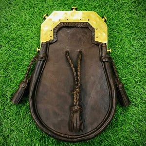 Puede incluir: Un sporran de cuero marrón oscuro con una pieza superior de latón pulido que presenta hendiduras circulares decorativas. La bolsa incluye tres borlas de cuero trenzado que cuelgan del frente y los lados, aseguradas por un marco de latón a juego.