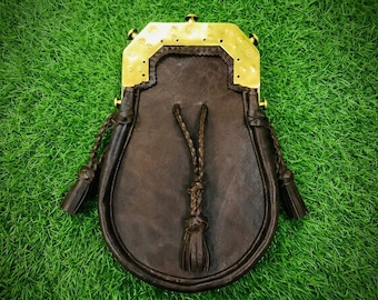 May include: A dark brown leather sporran featuring a polished brass cantle with decorative circular indentations. The pouch includes three braided leather tassels hanging from the front and sides, secured by a matching brass frame and hardware.