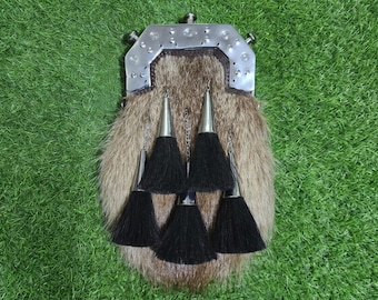 May include: A sporran featuring a brown fur body with a silver-toned metal cantle at the top. The cantle is decorated with small circular studs. Three black tassels with silver-toned conical caps hang from the front, suspended by thin chains.