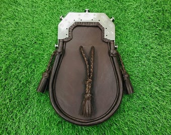 Può includere: Uno sporran in pelle marrone con una chiusura in metallo color argento lucido, decorata con incisioni e borchie. La borsa presenta tre cordini in pelle intrecciata con nappe abbinate che pendono dal davanti e dai lati, fissati da una cornice metallica.