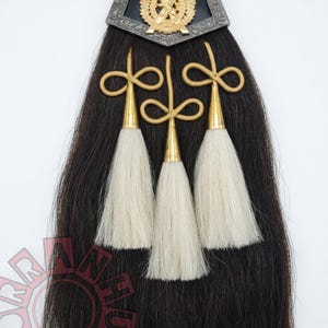 May include: A traditional Scottish sporran, featuring a black horsehair body, a silver-toned metal top with a gold emblem, and three white tassels. Gold-colored accents are also visible.