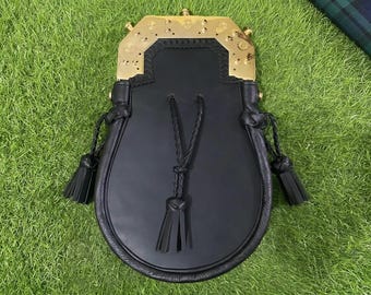 Könnte beinhalten: Ein schwarzer Ledersporran mit einem polierten, goldfarbenen Metallbeschlag, der mit dekorativen Nieten versehen ist. Die Tasche verfügt über drei schwarze Lederquasten an geflochtenen Kordeln sowie eine glatte Front aus schwarzem Leder.