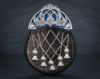 Puede incluir: Un sporran con cuerpo de piel marrón oscuro y borde de cuero negro. La solapa superior está adornada con una placa de metal plateado con incrustaciones de esmalte azul y un motivo de cardo. Tres cadenas plateadas cuelgan con cinco borlas.