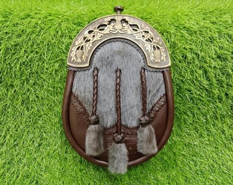 May include: A sporran featuring a dark brown leather body and a central panel of grey fur. The top is adorned with an ornate brass-colored metal plate displaying thistle motifs. Three braided leather tassels with fur tips hang from the front.