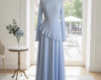 May include: A light blue, long-sleeved dress with a ruffled accent on the waist, displayed on a mannequin. The dress has a flowing skirt and is set in front of a large window. The dress is labeled "Etsy Exclusive".