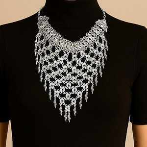 May include: Silver-toned statement necklace with intricate openwork design. The necklace features a bib-style front with a geometric pattern and dangling fringe. The jewelry is worn against a black background, highlighting the metallic shine and detailed craftsmanship.