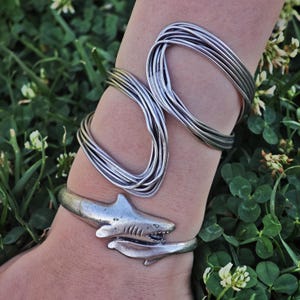 May include: Silver-toned metal bracelets. One bracelet features a shark design, wrapping around the wrist. The other bracelet is made of multiple strands of silver wire, forming a loop design. The bracelets are worn on a wrist.