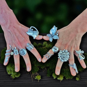 May include: A collection of silver-toned rings with various designs, including a cat, butterfly, snake, and sunflower. The rings are displayed on a bed of green moss, showcasing the intricate details and craftsmanship of each piece.