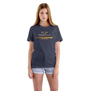 Puede incluir: Camiseta azul marino con el texto dorado "NOT NOW I'M HYPERFOCUSING". La camiseta tiene una barra horizontal dorada. La modelo lleva pantalones cortos vaqueros.