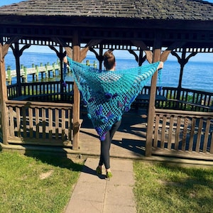 May include: A turquoise crocheted shawl with purple accents is displayed outdoors. The shawl is held open, showcasing its intricate net-like design. The background includes a wooden gazebo, water, and a clear blue sky.