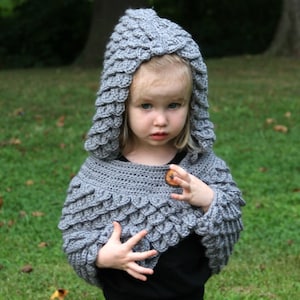 Può includere: Una cappa con cappuccio in crochet grigio con un motivo a scaglie. La cappa ha una chiusura a bottone ed è indossata da un bambino.