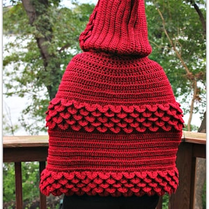 May include: A red crocheted hooded cape with a scalloped edge. The cape is made with a textured stitch and has a deep hood.