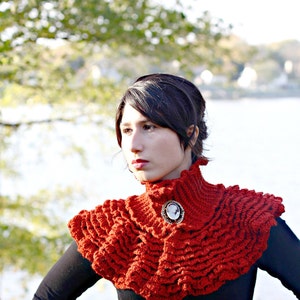 May include: A woman wearing a red crocheted cowl with a ruffled edge and a cameo brooch. The cowl is worn over a black mock neck shirt.