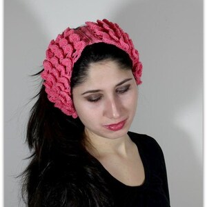 May include: A pink crocheted headband with a flower design. The headband is worn by a person with long dark hair.