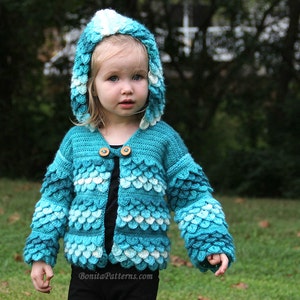 May include: A young child wearing a blue and white crocheted hooded cardigan with a scale pattern. The cardigan has a button closure and is made with a soft yarn.