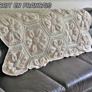 May include: A white crocheted throw blanket with a floral pattern. The blanket is folded and placed on a brown leather couch.
