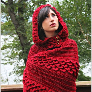 May include: A red crocheted hooded shawl with a textured, scale-like pattern. The shawl is worn by a person with dark hair and is styled with the hood up.