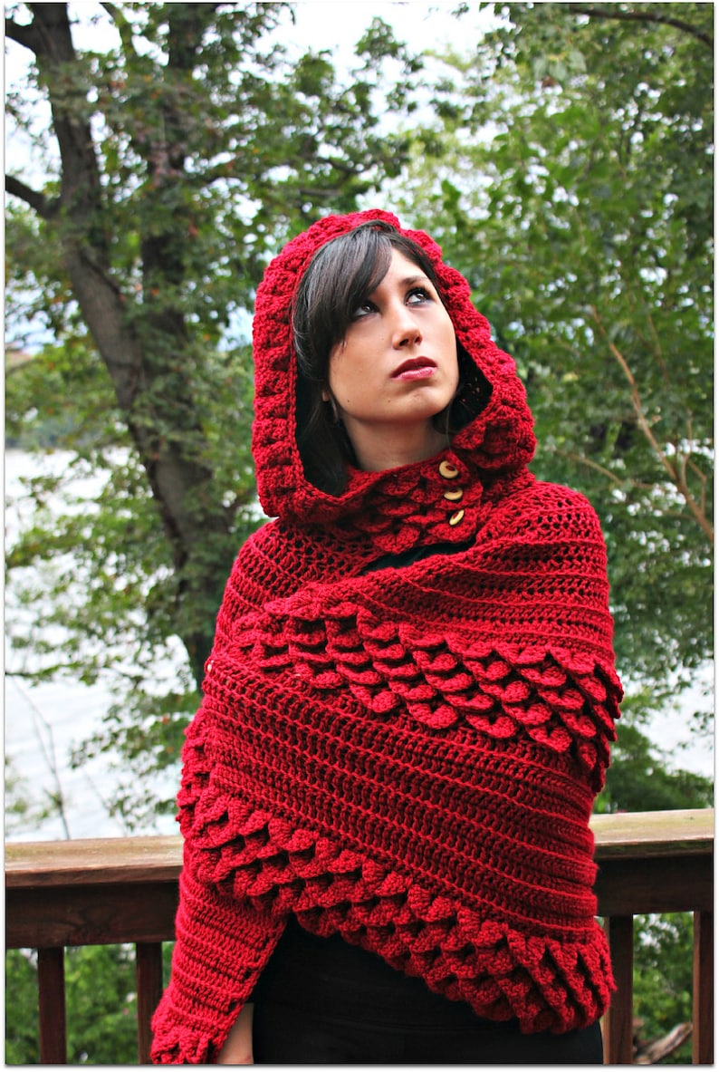 May include: A red crocheted hooded shawl with a textured, scale-like pattern. The shawl is worn by a person with dark hair and is styled with the hood up.