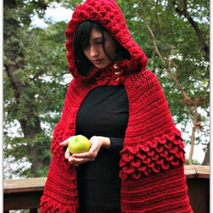 May include: A red crocheted hooded cape with a scalloped edge and a green apple held in the hand of the person wearing the cape.