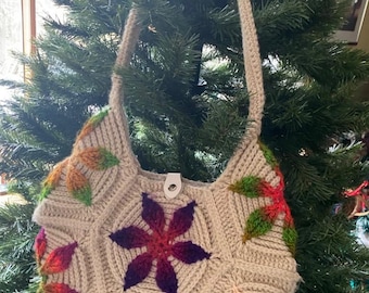 May include: A crocheted beige shoulder bag with a floral design. The bag features a purple, red, and orange flower in the center and green leaves around the edges.