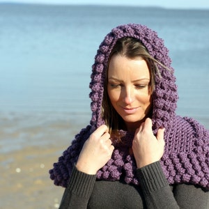 May include: A purple crocheted hooded cowl with a textured bobble stitch. The cowl is worn by a person standing in front of a beach.