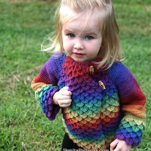 Puede incluir: Un niño pequeño lleva un suéter de crochet arcoíris con un patrón de escamas. El suéter tiene botones de madera y es una mezcla vibrante de colores.
