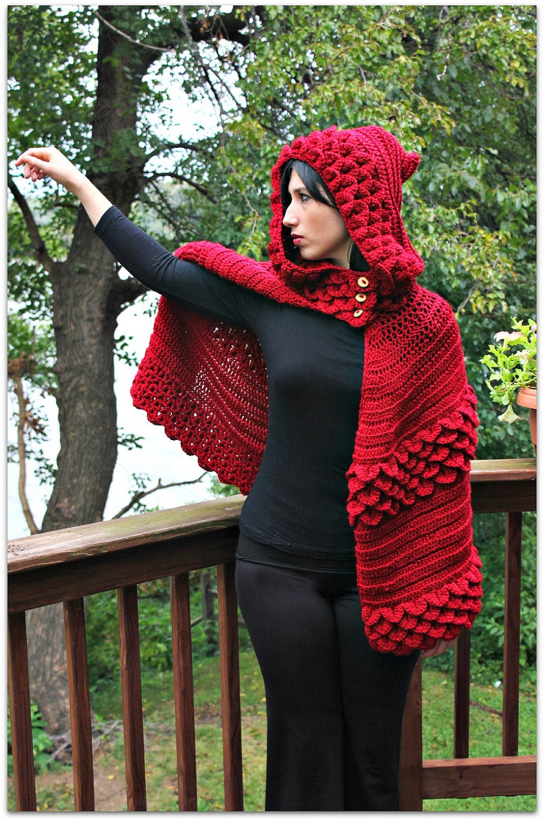 May include: A red crocheted hooded cape with a textured design. The cape has a scalloped edge and is worn over a black top and pants.