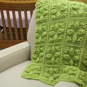 Puede incluir: Una manta de bebé de ganchillo verde con un patrón floral. La manta está colocada sobre un sillón beige.