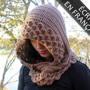 Könnte beinhalten: Ein brauner, gehäkelter Kapuzen-Schal mit einem strukturierten Muster. Der Schal wird von einer Person mit langen, dunklen Haaren getragen. Der Text "ÉCRIT EN FRANÇAIS" ist in der oberen rechten Ecke des Bildes sichtbar.