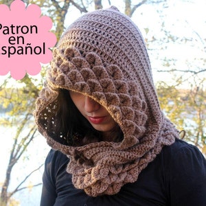 May include: A brown crocheted hooded cowl with a textured pattern. The cowl is worn by a person with long dark hair. The text "Patron en Español" is visible in the image.