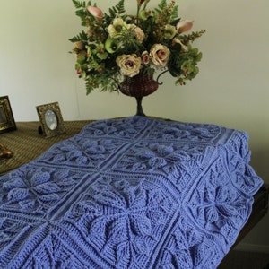 Puede incluir: Una manta de ganchillo morada con un patrón texturizado de flores. La manta está doblada y drapeada sobre una mesa con un jarrón de flores en la parte superior.