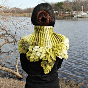 May include: A green and white crocheted cowl with a leaf design. The cowl is worn by a person standing in front of a body of water.