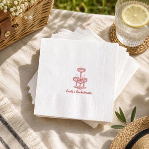 May include: A stack of square white paper napkins featuring a red line art illustration of a champagne tower and the text "Emily's Bachelorette" printed in a red script font centered on the front-most napkin in the stack.