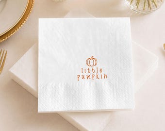 May include: A square white paper napkin featuring a small orange line drawing of a pumpkin centered above the text "little pumpkin" in a simple orange font. The napkin has a textured border and is stacked on top of other identical white napkins.