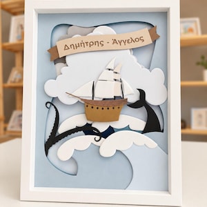 May include: A framed 3D paper art piece featuring a brown sailing ship on light blue waves with white foam. A black tentacle and whale tail emerge from the water. A tan banner at the top displays the Greek text "Δημήτρης - Άγγελος" in dark brown.