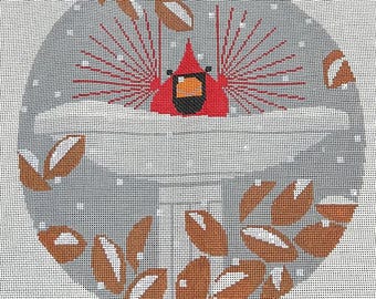 May include: A round needlepoint design featuring a red cardinal bird with a black beak and orange accents, set against a gray background with brown leaves. The design is surrounded by a white border and small white dots.