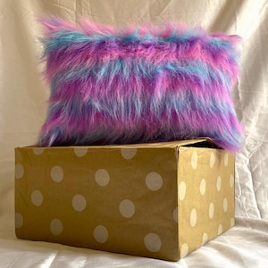 May include: A rectangular, fluffy pillow in shades of purple, blue, and pink rests on a brown box with white polka dots. The pillow has long, shaggy fur, creating a soft and textured appearance. The box is made of cardboard.