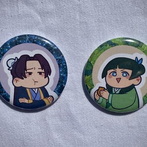 Può includere: Due spille rotonde con illustrazioni in stile anime. Una presenta un personaggio in blu e oro con uno sfondo stellato. L'altra mostra un personaggio in verde con un bordo di foglie, che tiene in mano un piccolo oggetto.