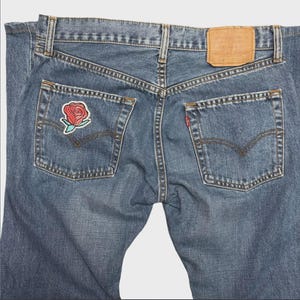 May include: Blue denim jeans with a rose patch on the back pocket. The jeans feature classic five-pocket styling, a leather patch, and a red Levi's tag. The jeans are a medium wash and show signs of wear.