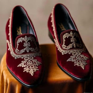 以下が含まれることがあります： 甲と側面に複雑なゴールドの刺繍が施された、深みのある赤のベルベットローファー。つま先が尖ったシルエットで、内側は黒い裏地、かかとは低い黒色のヒールになっています。