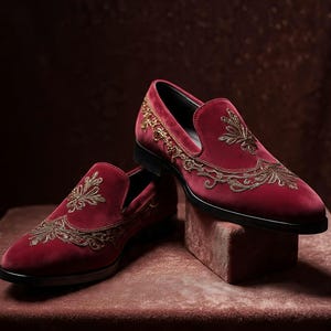 May include: A pair of burgundy velvet loafers with gold embroidered floral designs. The shoes have a classic slip-on style with a black sole and are displayed on a matching burgundy surface.