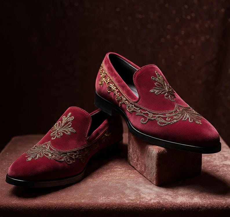 May include: A pair of burgundy velvet loafers with gold embroidered floral designs. The shoes have a classic slip-on style with a black sole and are displayed on a matching burgundy surface.